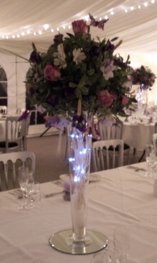 Ball shaped arrangement including roses, freesia and lizianthus on top of conical vase with twig lights