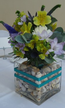 Yellow alstromeria, lilac freesia, purple veronica, white gerbera arranged in tank vase with shells and ribbon